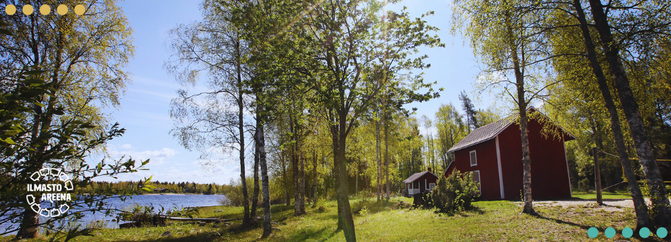 Kesäinen ja aurinkoinen mökkimaisema. Oikealla näkyy kaksi punaista puurakennusta, vasemmalla vesistöä. Pihalla kasvaa lehtipuita.