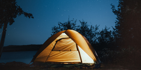 Sisältä valaistu teltta yön pimeydessä rannan äärellä. Taustalla näkyy tähtiä.