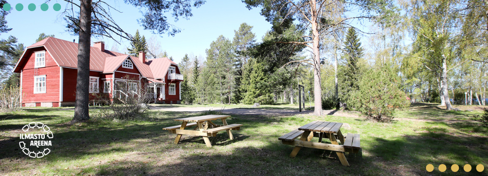 Karhun leirikeskuksen pihapiiri aurinkoisena kesäpäivänä. Taustalla näkyy suuri punainen rakennus, etualalla muutama puinen pöytä ja penkki.